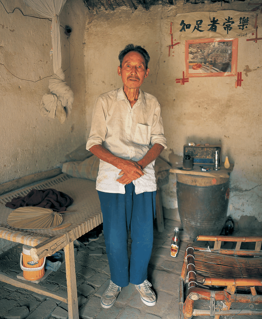 一位老人在简陋的土房中站立，墙上写着知足者常乐