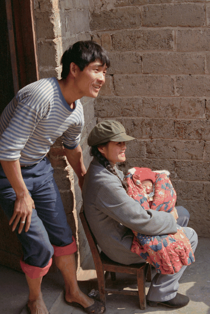 一张充满年代感的家庭合影，年轻父母与婴儿在砖墙前微笑