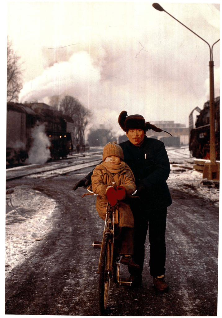老照片：父亲推着自行车，车座上的孩子与背景中的蒸汽机车